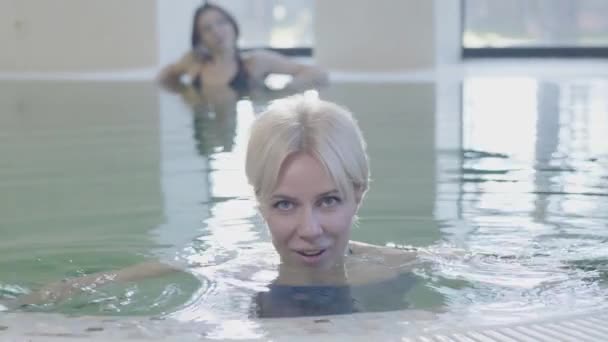 Close-up of positive blond grey-eyed woman posing at poolside with relaxed brunette girl resting at the background. Portrait of charming Caucasian lady enjoying vacations in luxurious hotel. - Materiał filmowy, wideo