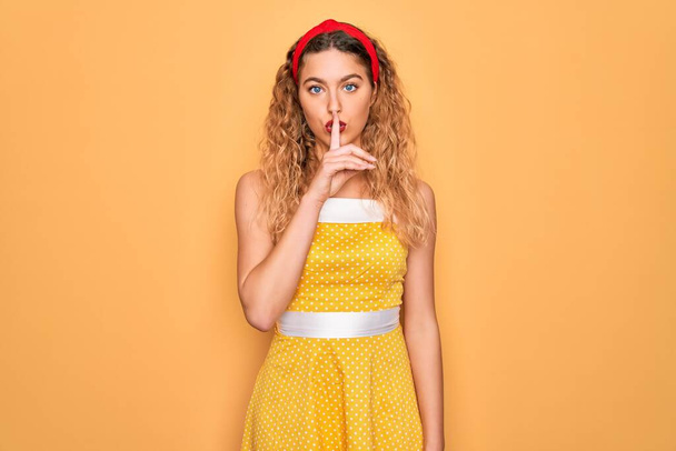 Beautiful blonde pin-up woman with blue eyes wearing diadem standing over yellow background asking to be quiet with finger on lips. Silence and secret concept. - Photo, Image