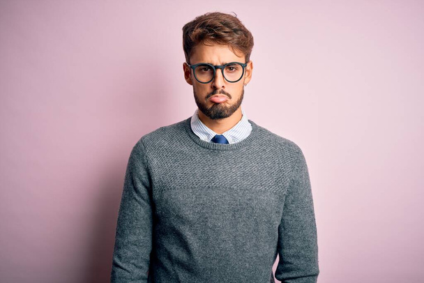 Νεαρός όμορφος άντρας με γενειάδα που φοράει γυαλιά και πουλόβερ στέκεται πάνω από ροζ φόντο μελαγχολικός και ανησυχεί για την αγωνία, κλαίει θυμωμένος και φοβισμένος. Θλιβερή έκφραση. - Φωτογραφία, εικόνα