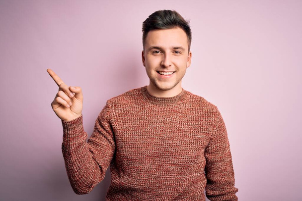 Joven hombre caucásico guapo usando suéter de invierno casual sobre fondo aislado rosa con una gran sonrisa en la cara, señalando con el dedo de la mano al lado mirando a la cámara
. - Foto, Imagen