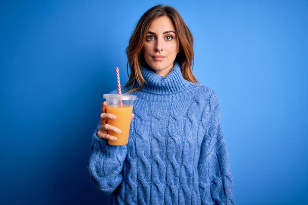 Jeune belle femme brune buvant du jus d'orange sain sur fond bleu avec une expression confiante sur la pensée intelligente du visage grave
 - Photo, image