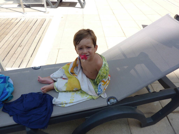 Маленький ребенок с пустышкой надевает солнцезащитные очки, сидя на шезлонге
 - Фото, изображение