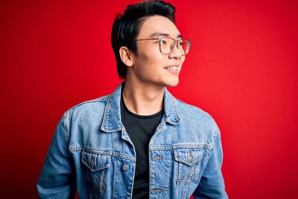 Молодой красивый китайский мужчина в джинсовой куртке и очках на красном фоне смотрит в сторону с улыбкой на лице, естественным выражением лица. Смеясь уверенно
. - Фото, изображение