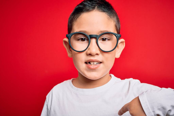 Mladý malý chytrý chlapec kluk nosí nerd brýle přes červené izolované pozadí s překvapivou tváří ukazující prstem na sebe - Fotografie, Obrázek
