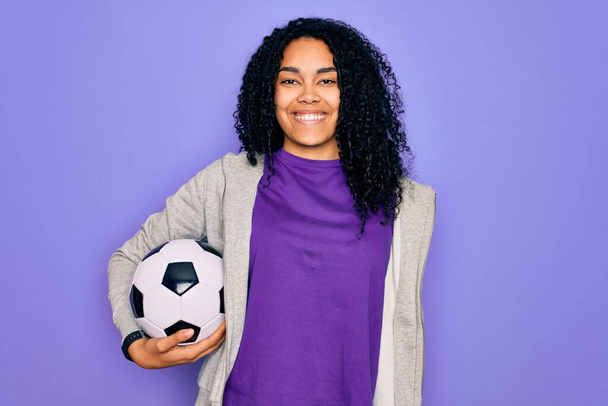 Afrykański amerykański kręcone gracz kobieta gra w piłkę nożną trzymając piłkę nożną na fioletowym tle ze szczęśliwą twarzą stojąc i uśmiechając się z pewnym uśmiechem pokazując zęby - Zdjęcie, obraz