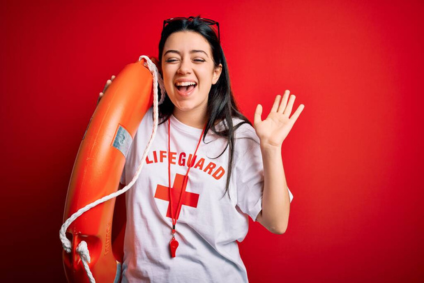 Young lifeguard woman wearing secury guard equipent holding rescue float over red background very happy and excited, winner expression celebrating victory screaming with big smile and raised hands - Photo, Image