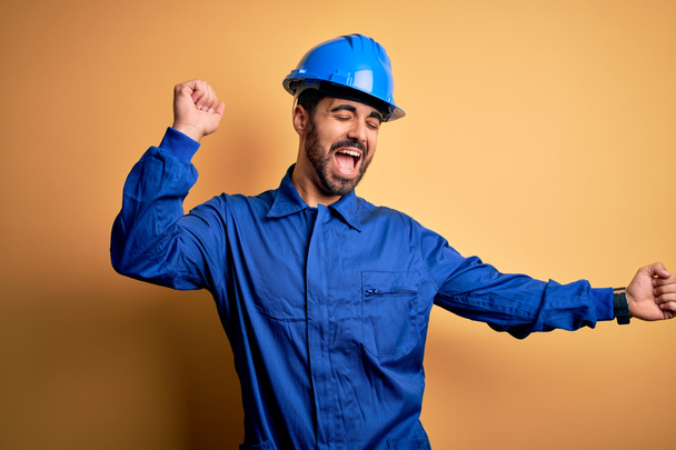Mechanic man with beard wearing blue uniform and safety helmet over yellow background Dancing happy and cheerful, smiling moving casual and confident listening to music - Photo, Image