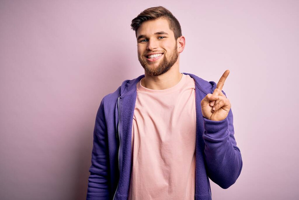 Молодий блондин з бородою і блакитними очима носить фіолетовий светр на рожевому фоні, показуючи і вказуючи пальцем номер один, посміхаючись впевнено і щасливо
. - Фото, зображення
