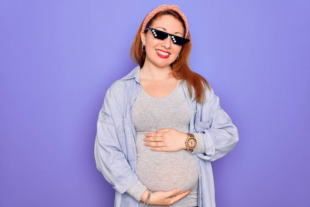 Jonge roodharige zwangere vrouw in verwachting van een baby met een grappige gangster zonnebril met een vrolijke en coole glimlach op haar gezicht. Geluksvogel. - Foto, afbeelding