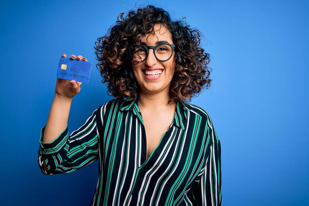 Giovane bella donna araba riccia con gli occhiali in possesso di soldi della carta di credito per il pagamento con una faccia felice in piedi e sorridente con un sorriso fiducioso che mostra i denti
 - Foto, immagini