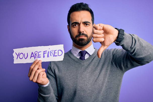 Young handsome businessman with beard holding paper with you are fired message with angry face, negative sign showing dislike with thumbs down, rejection concept - Photo, Image