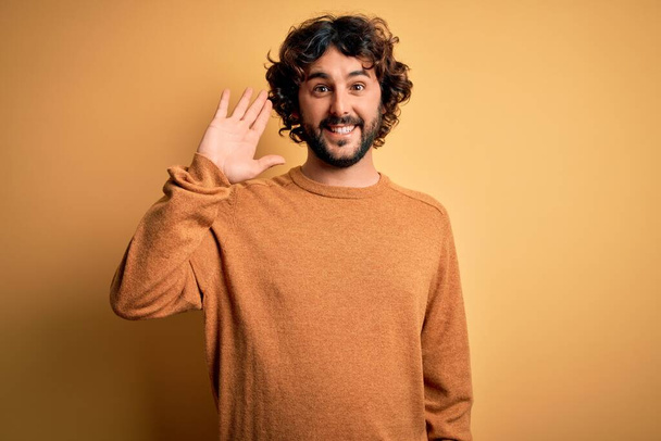 Junger gutaussehender Mann mit Bart in lässigem Pullover vor gelbem Hintergrund, der fröhlich grüßt und lächelt, freundliche Willkommensgeste - Foto, Bild