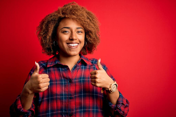 Junge schöne afroamerikanische Afro-Frau mit lockigem Haar trägt lässiges Hemd Erfolgszeichen tun positive Geste mit der Hand, Daumen nach oben lächelnd und glücklich. Fröhliche Miene und Siegergeste. - Foto, Bild