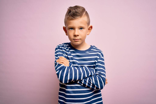 Nuori valkoihoinen poika sinisilmäinen yllään meripeninkulman raidallinen paita vaaleanpunainen tausta skeptinen ja hermostunut, paheksuva ilme kasvot ristissä kädet. Kielteinen henkilö
. - Valokuva, kuva