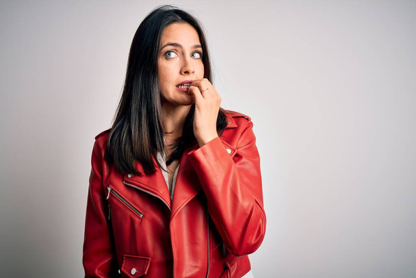 Young brunette woman with blue eyes wearing red casual jacket over white background looking stressed and nervous with hands on mouth biting nails. Anxiety problem. - Fotó, kép