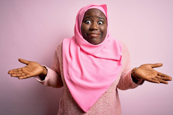 Fiatal afro-amerikai plusz méretű nő visel muszlim hidzsáb felett elszigetelt rózsaszín háttér tudatlan és zavaros kifejezés karok és kezek felemelt. Kétséges fogalom. - Fotó, kép