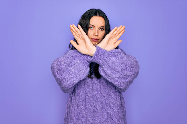 Молодая красивая женщина в свитере в водолазке стоит на фиолетовом фоне Отвергающее выражение, скрещивающее руки и ладони, делающее негативный знак, злое лицо
 - Фото, изображение