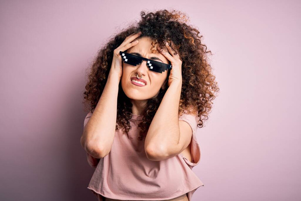 Young beautiful woman with curly hair and piercing wearing funny thug life sunglasses suffering from headache desperate and stressed because pain and migraine. Hands on head. - Foto, imagen