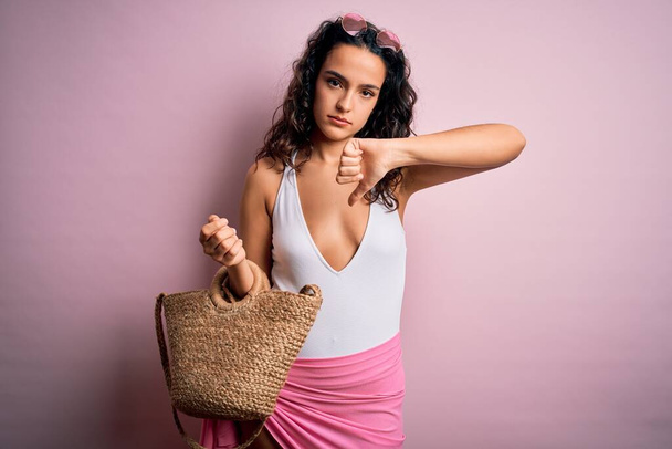 Молодая красивая женщина с вьющимися волосами на отдыхе в купальнике, держащая пляжную сумку с сердитым лицом, негативный знак, показывающий неприязнь с опущенными пальцами, концепция отказа
 - Фото, изображение