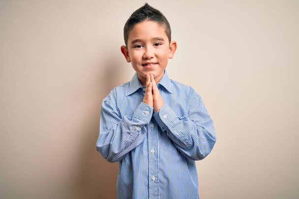 Маленький мальчик в элегантной рубашке, стоя на изолированном фоне, молится руками, прося прощения, уверенно улыбаясь.
. - Фото, изображение