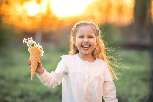 Ένα όμορφο κοριτσάκι γελάει και κρατάει τα ανοιξιάτικα λουλούδια του δέντρου σε ένα χωνάκι βάφλας στο χέρι της. Η ιδέα είναι η αρχή μιας νέας ανοιξιάτικης διάθεσης και παιδιάστικου αυθορμητισμού. Ζεστό ηλιοβασίλεμα. - Φωτογραφία, εικόνα