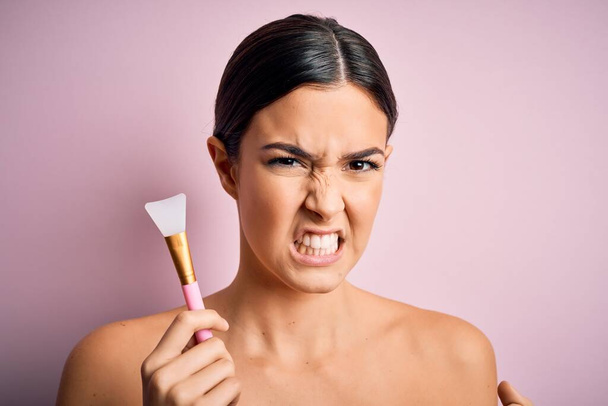 Young beautiful girl applying cosmetic using spatula over pink background annoyed and frustrated shouting with anger, crazy and yelling with raised hand, anger concept - Photo, Image