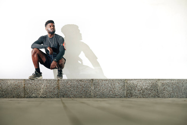 Seriöser, selbstbewusster junger schwarzer Sportler sitzt auf Bordstein - Foto, Bild