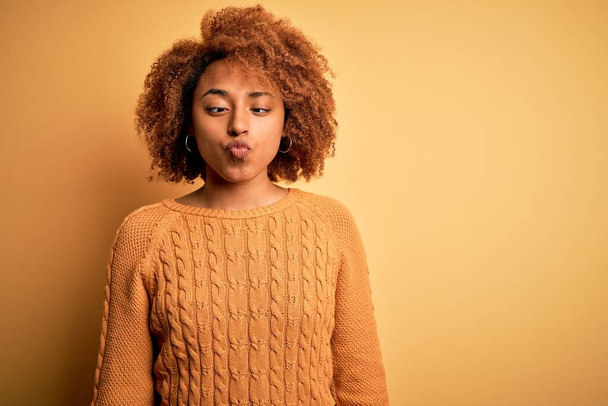 Молодая красивая афро-американка с кудрявыми волосами в повседневном свитере делает рыбное лицо с губами, сумасшедший и смешной жест. Смешное выражение
. - Фото, изображение