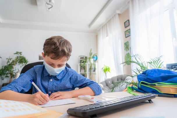 Boy in face mask with computer does his homework during a coronavirus quarantine. concept of online education. Social distance, self- isolation. Distance learning due to virus, flu, epidemic COVID-19 - Foto, Bild
