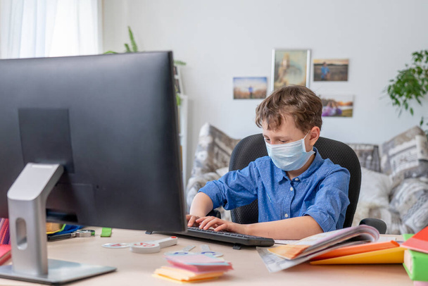 boy with a face mask does his homework using a computer during a coronavirus quarantine. the concept of online education. Social distance, self-isolation. Distance learning due to virus, flu, epidemic COVID-19 - Foto, Imagem