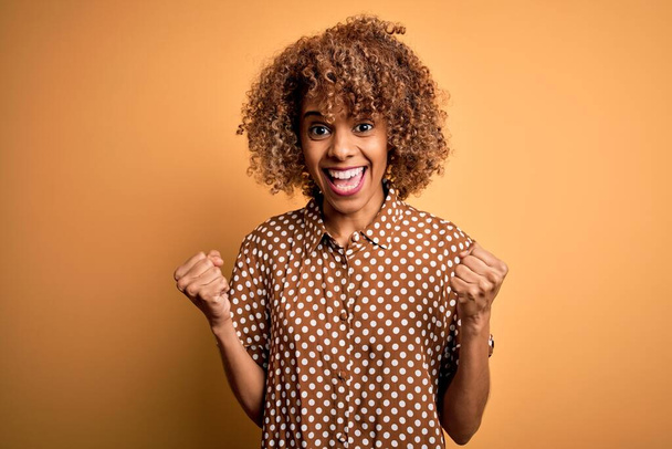 Sarı arka plan üzerinde gündelik tişört giyen genç ve güzel Afro-Amerikalı kadın kollarını kaldırarak ve açık gözlerle başarıyı kutluyor ve şaşırıyor. Kazanan konsept. - Fotoğraf, Görsel