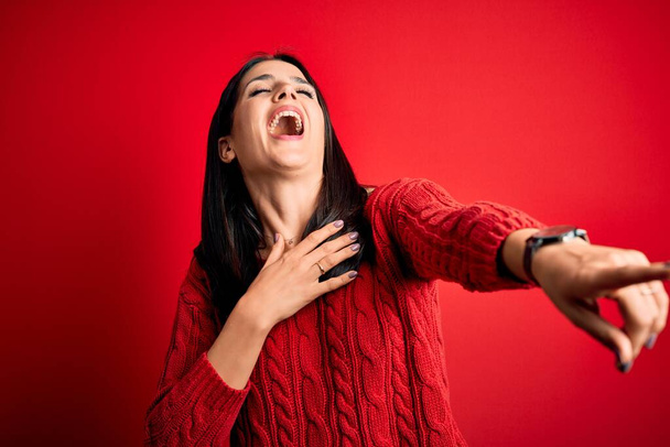 Giovane donna bruna con gli occhi blu che indossa maglione casual su sfondo rosso isolato ridendo di te, indicando il dito alla fotocamera con mano sul corpo, espressione di vergogna
 - Foto, immagini