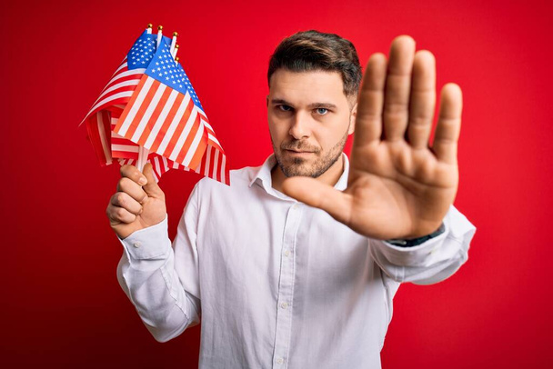 Mladý muž s modrýma očima drží vlajku Spojených států amerických nad červeným izolovaným pozadím s otevřenou rukou dělá stopku s vážným a sebevědomým výrazem, obranné gesto - Fotografie, Obrázek