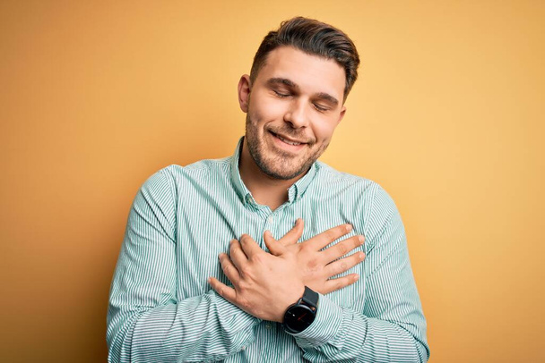 Young business man with blue eyes wearing elegant green shirt over yellow background smiling with hands on chest with closed eyes and grateful gesture on face. Health concept. - Photo, Image