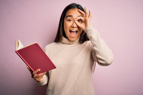 Joven asiático inteligente mujer sosteniendo libro diario de pie sobre aislado rosa fondo con feliz cara sonriendo haciendo ok signo con la mano en el ojo mirando a través de los dedos
 - Foto, Imagen