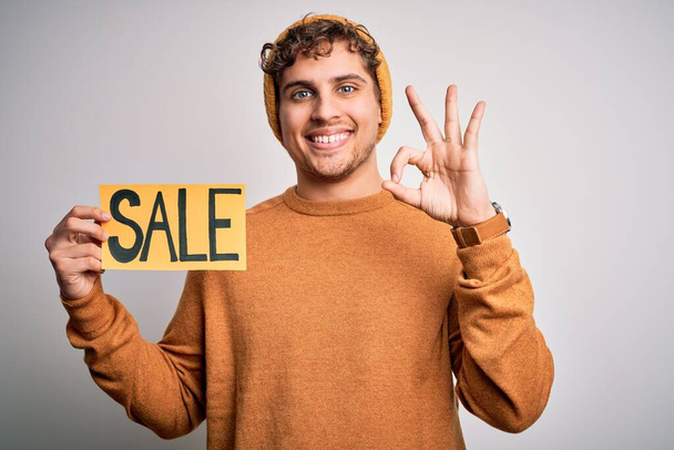 Jonge blonde man met krullend haar houden verkoop poster staan over geïsoleerde witte achtergrond doet ok teken met vingers, uitstekend symbool - Foto, afbeelding
