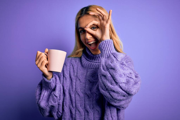 Nuori kaunis blondi nainen juo kupin kahvia yli eristetty violetti tausta onnellinen kasvot hymyillen tekee ok merkki käsin silmään katsellen sormien läpi
 - Valokuva, kuva