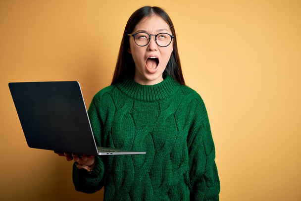 Young asian business woman wearing glasses and working using computer laptop angry and mad screaming frustrated and furious, shouting with anger. Rage and aggressive concept. - 写真・画像