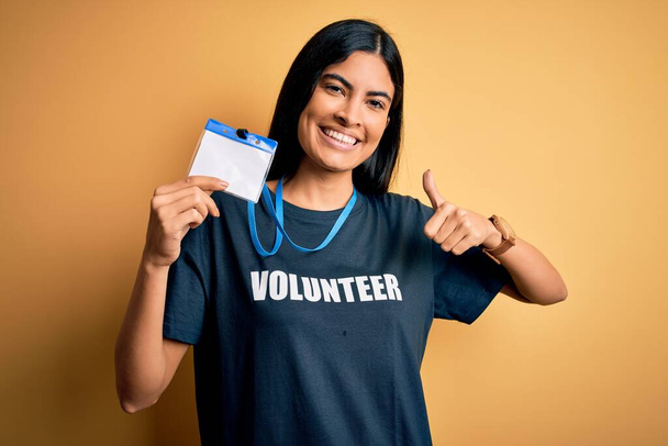Young beautiful hispanic woman wearing volunteer t-shirt and showing identification happy with big smile doing ok sign, thumb up with fingers, excellent sign - Fotó, kép