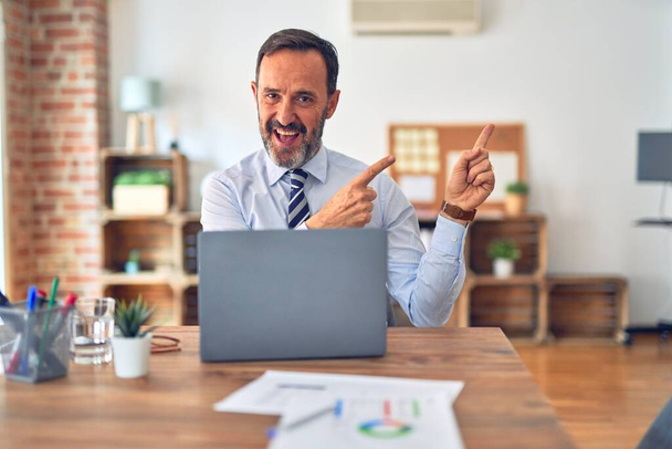Homem de negócios bonito de meia idade usando gravata sentado usando laptop no escritório sorrindo e olhando para a câmera apontando com duas mãos e dedos para o lado
. - Foto, Imagem