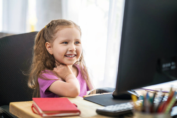 little girl uses a computer to study at home. The child smiles happily and gets knowledge remotely. digital concept of e-learning, during epidemic, quarantine and self-isolation. Remote e-learning. - Фото, зображення