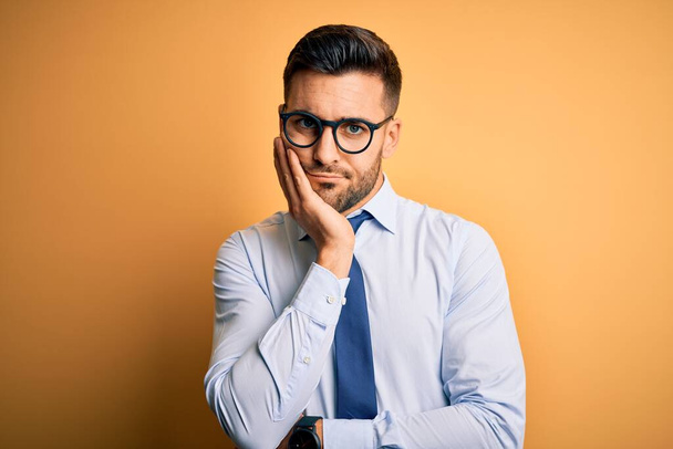 Young handsome businessman wearing tie and glasses standing over yellow background thinking looking tired and bored with depression problems with crossed arms. - Photo, Image