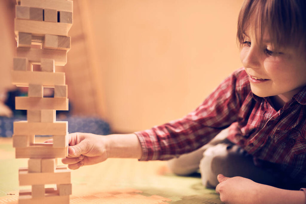 Preschool cute boy playing in a table game with wooden blocks at home. Players take turns removing one block at a time from a tower. Each block removed is then placed on top of the tower. - Fotografie, Obrázek