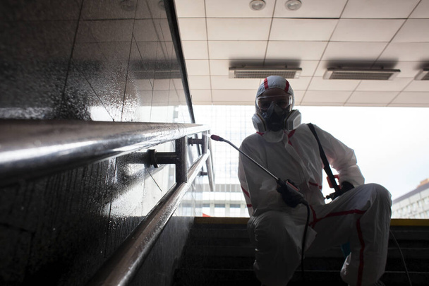 desinfectie van de stad van het coronavirus, een man in een beschermend pak en ademhalingsapparaat reinigt de leuningen van de trap met een chemische spray, een sanitair werker, chemische behandeling - Foto, afbeelding