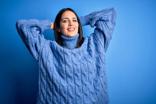 Young brunette woman with blue eyes wearing casual turtleneck sweater relaxing and stretching, arms and hands behind head and neck smiling happy - Photo, Image
