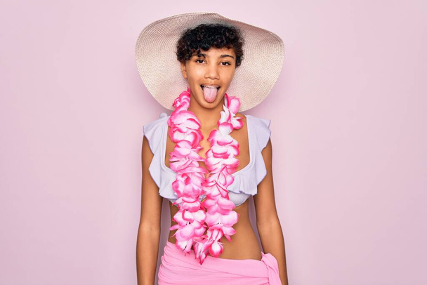 Młoda, piękna afrykańska turystka ubrana w bikini i hawajskie kwiaty lei wystające z języczkiem zadowolona ze śmiesznej ekspresji. Koncepcja emocji. - Zdjęcie, obraz