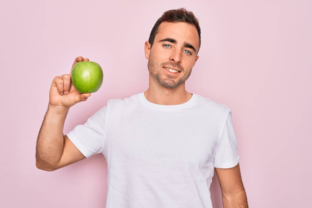 Young handsome man with blue eyes eating healthy green apple over isolated pink background with a happy face standing and smiling with a confident smile showing teeth - 写真・画像