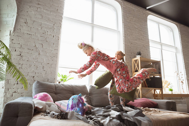Quiet little girls playing in a bedroom in cute pajamas, home style and comfort, laughting and fighting pillows, jumping high - Foto, Imagem