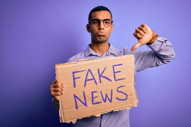 Jonge knappe Afro-Amerikaanse man protesteert met spandoek met nep nieuwsbericht met boos gezicht, negatief teken toont afkeer met duimen naar beneden, afwijzing concept - Foto, afbeelding