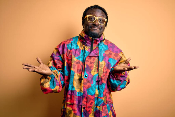 Handsome african american man wearing colorful coat and glasses over yellow background clueless and confused expression with arms and hands raised. Doubt concept. - Photo, Image